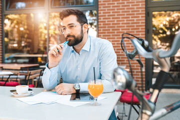 Young thoughtful man with glasses and beard thinking how to improve his future business. He is surrounded by papers and digital equipment.