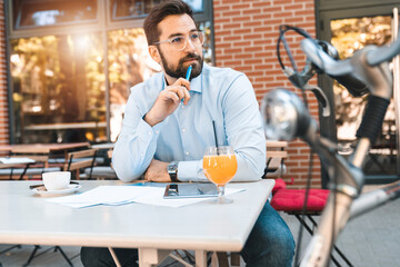 Young thoughtful businessman with glasses and beard holding pen, looking aside and thinking how to...
