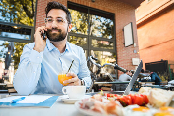 Cheerful businessman in front of table with laptop and paper document on it in restaurant, smiling and holding glass. Food is served on table.