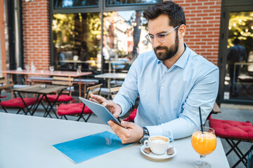 Dedicated businessman sitting outdoors and having a coffee break. He is using his digital tablet while waiting his coffee to cool down.