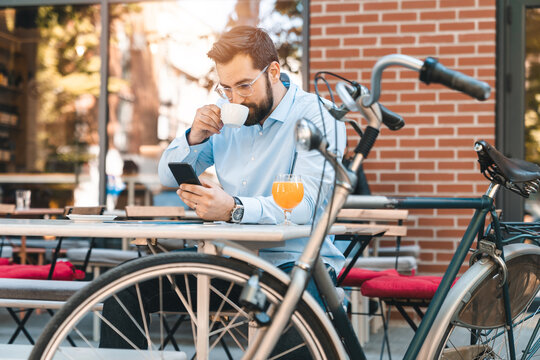 Focused businessman in his 30s drinking coffee outdoors and texting on his smartphone during the late afternoon with a parked bicycle next to the table.