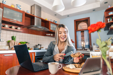 Cheerful blonde woman sitting at table and eating breakfast in kitchen. Smiling girl is holding...