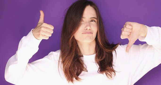 Young woman in white pullover making good-bad sign isolated on purple background. Girl make choice, or makes a decision thumb up or thumb down, like or dislike, yes or no. Thumb up win.