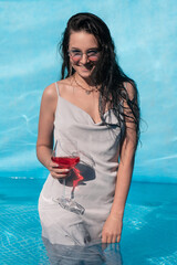 Beautiful girl with a glass of alcohol posing in the pool, summer vacation