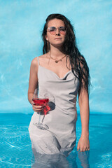 Beautiful girl with a glass of alcohol posing in the pool, summer vacation