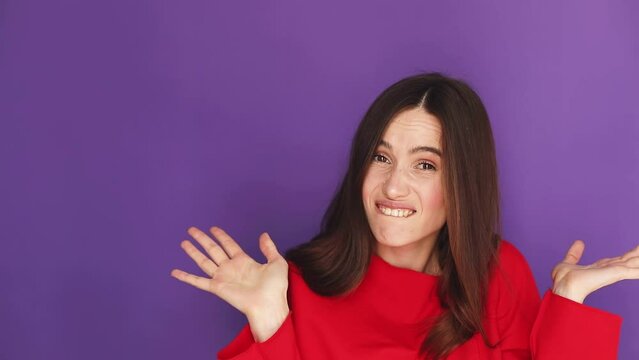 Guilty confused woman making oops, I'm sorry expression. Girl wear red sweater is ashamed and feeling uncomfortable because of guilt on violet studio background.
