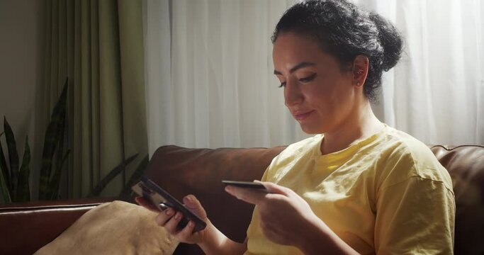 Smiling young woman customer holding credit card and smartphone sitting on couch at home.Happy female shopper with glasses using instant easy mobile payments making purchase in online store.Banking ap