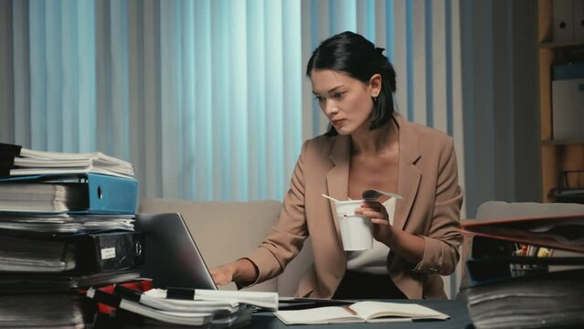 Medium shot of young female hungry office worker eating instant noodles while working on report sitting at office desk at night timeMedium shot of young female hungry office worker eating instant nood