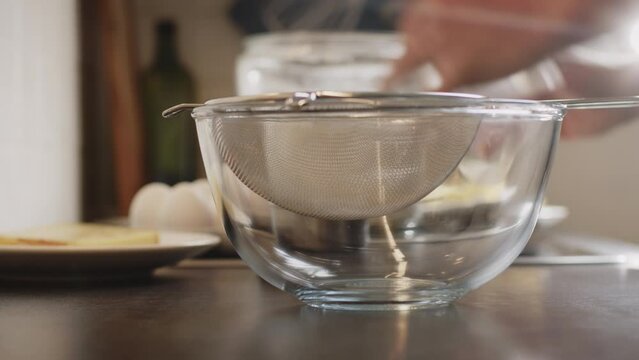 Home cooking - a man sieving flour in a transparent glass mixing bowl. Low angle closeup shot.
