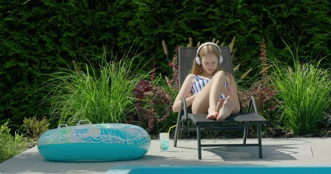 A teenage girl is resting in a sun lounger by the pool. Uses a smartphone, listens to music on headphones