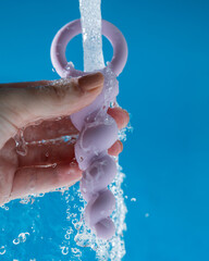 Woman holding lilac anal beads under running water on blue background. Sex toy hygiene concept.