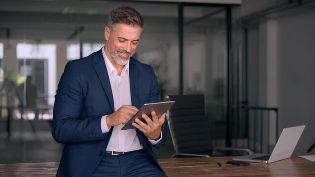 Happy Latin Hispanic bearded stylish mature adult professional business man, smiling Indian senior businessman CEO holding digital tablet using fintech tab application standing inside company office.