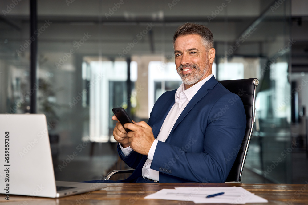 Canvas Prints Middle aged Hispanic business manager ceo using cell phone mobile app, looking at camera. Smiling Latin or Indian mature man businessman holding smartphone sit in office working online in office.