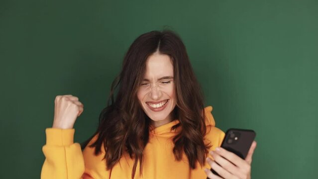 Extremely happy, satisfied girl in yellow hoody is using her smartphone isolated on green studio. Funny smily girl is texting messages, playing with her mobile phone. Good fortune. Win gesture.