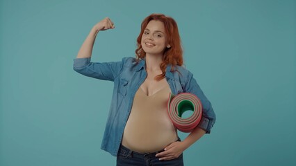 Portrait of a young pregnant woman holding a yoga mat in the studio on a blue background, showing a...