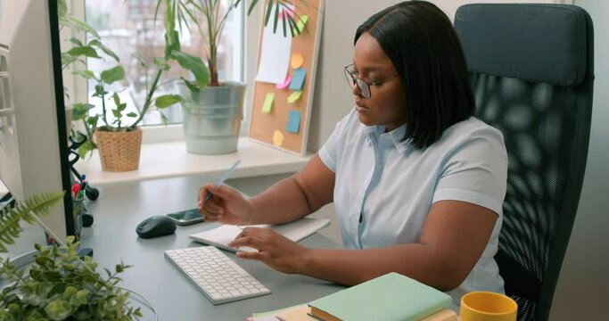 Young serious ambitious Black business woman sitting at table and writing in notebook Freelancer working, writing down new ideas, slow motion. planning agenda with personal planner and online calendar