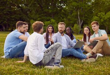 Naklejka premium Group of students friends preparing for classes in nature or discussing social media in summer park. Diverse people sitting on the green grass outdoor with laptop together and talking.