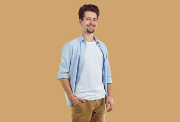 Studio portrait of a happy, smiling young man in casual clothes. Male model in a white T shirt, light blue shirt and brown pants standing with his hand in his pocket on a beige brown background