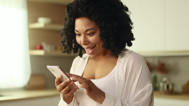 Portrait of young curly woman looking at smartphone cellphone screen with nod of approval and looking at camera pointing at the phone at home Advise Approve Recommend Choice concept