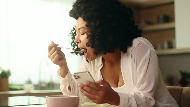 Smiling young curly woman enjoying beautiful morning with tasty breakfast hold smartphone watching online videos entertainment program in social media alone in kitchen at home