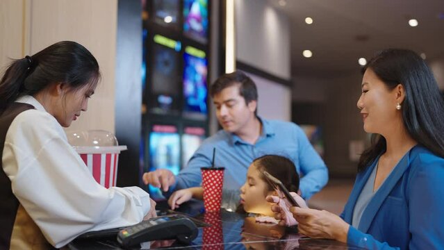 Happy Asian family choosing seats and buying movie ticket in cinema box office. Mother using smartphone scan for paying while buying movie ticket in cinema box office. Entertainment concept