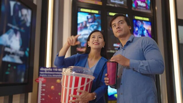 Happy Asian couple holding popcorn bucket while choosing movie at cinema box office. Asian couple having date at movie theater on weekend. Entertainment concept