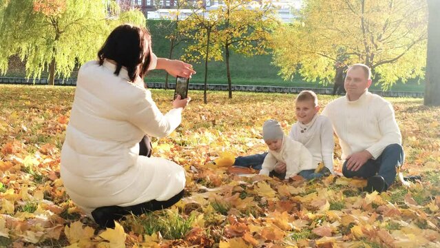 A happy family of four spend time in the autumn park. Mother takes a picture of her family on a mobile phone