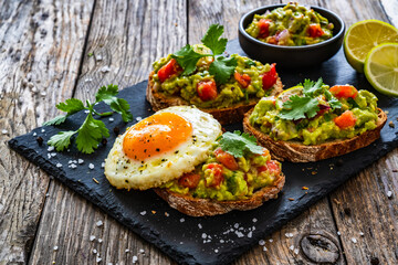 Tasty bruschetta with sunny side up egg, guacamole, tomato, and onion on wooden table
