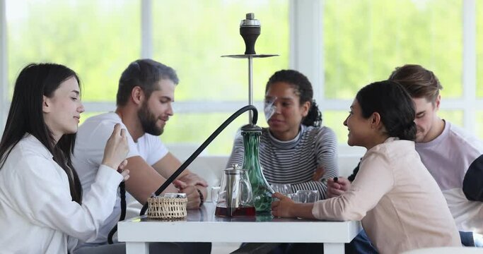 Positive relaxed young multiethnic friends smoking shisha in hookah lounge, talking, drinking tea, enjoying carefree conversation, smiling, laughing, resting on couches at table