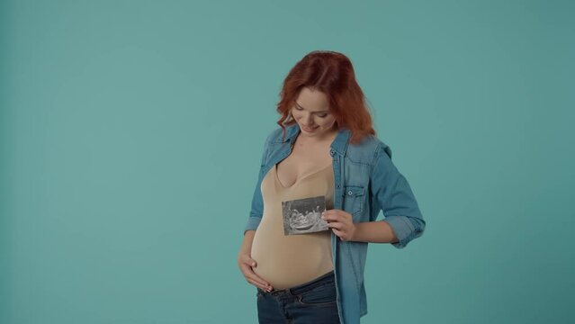 A pragnant woman holds an ultrasound scan and strokes her belly. Happy yong woman in the studio on a blue background. The concept of pregnancy, motherhood, tenderness, responsibility, growing up.