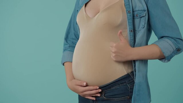 A pregnant woman strokes her bloated stomach with love and tenderness, shows a thumbs up. Bloated belly of a pregnant woman in the studio on a blue background close up.