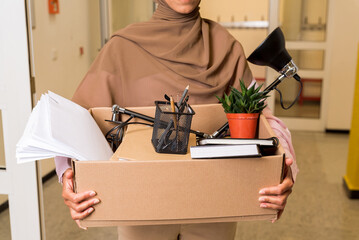 Elegant Muslim woman in hijab holding a box with personal items in the office going through the doors to a new job or getting fired. Coworking space and employee concept with personal items for work