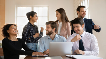 Overjoyed young multiethnic employees sit at desk in office laugh work together on laptop at meeting, happy smiling diverse colleagues cooperate on computer, team have fun collaborating at briefing
