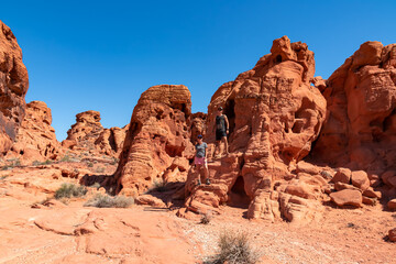 Fototapeta premium Couple at the entrance of windstone arch (fire cave) in Valley of Fire State Park, Mojave desert, Nevada, USA. Scenic view of beehive shaped red sandstone rock formations. Barren deserted landscape