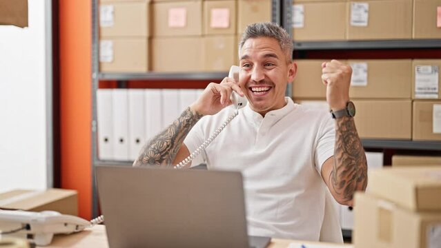 Young hispanic man ecommerce business worker using laptop talking on telephone celebrating at office