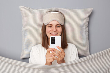 Cheerful smiling young woman wearing white T-shirt and sleep eye mask lies under blanket in bedroom...
