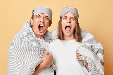 Angry couple man and woman in sleep eye mask wrapped in blanket isolated over beige background...