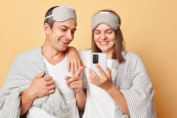 Happy smiling couple man and woman in sleep eye mask wrapped in blanket isolated over beige...