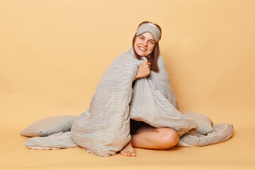Cute joyful satisfied woman wearing pajama and blindfold sitting on floor wrapped in blanket...