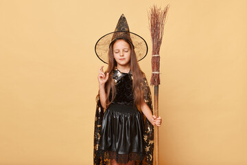 Hopeful little girl with long hair black cloak clothing with broom isolated over beige background standing with closed eyes and crossed fingers dreaming magic spell worked