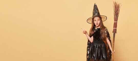 Childhood fascination with magic. Surprised little girl with long hair black cloak clothing with broom isolated over beige background kid pointing away at copy space for promotion