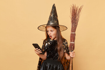 Shocked little girl with long hair black cloak clothing with broom isolated over beige background...