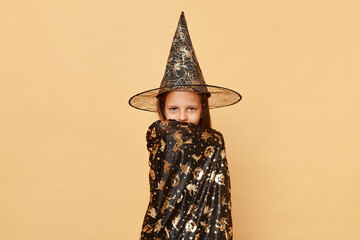 Beautiful little girl wearing witch costume and carnival cone hat isolated over beige background...