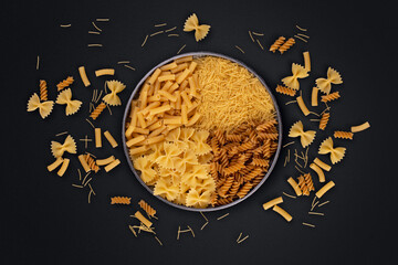 Variety of types and shapes of dry pasta in bowl, top view.