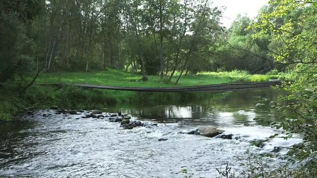 static wide shot of footbridge over a forest river with stones and small rapids