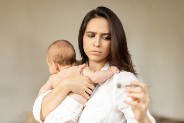 Concerned mother looking at electronic thermometer, holding her sick little kid, worried mom with...