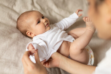 Mother dressing up her cute little baby after waking up, putting bodysuit, kid lying on bed at home, above view. Mother and child concept
