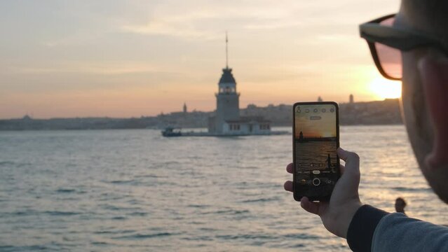 Man traveler photographing Maiden's Tower in Bosphorus strait in Istanbul at sunset using camera on his mobile phone. Caucasian guy tourist taking photo on smartphone