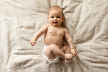 Cute little baby lying on bed, looking at camera and smiling, infant kid playing with his toes while resting in bedroom, top view, copy space