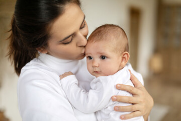 Caring young mom holding and hugging her adorable baby, enjoying motherhood and child care, standing in bedroom at home. Mother's day concept